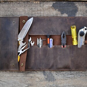 May include: A brown leather tool roll with multiple pockets and slots for holding tools. The roll is open and contains a variety of tools, including a knife, pliers, screwdrivers, and a utility knife.