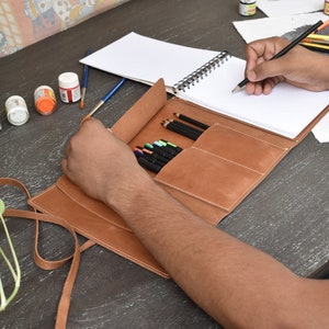 May include: A tan leather roll-up organizer featuring multiple interior pockets for pencils and pens, accompanied by a spiral-bound sketchbook with blank white pages. The organizer includes a long leather strap for securing the folded case.
