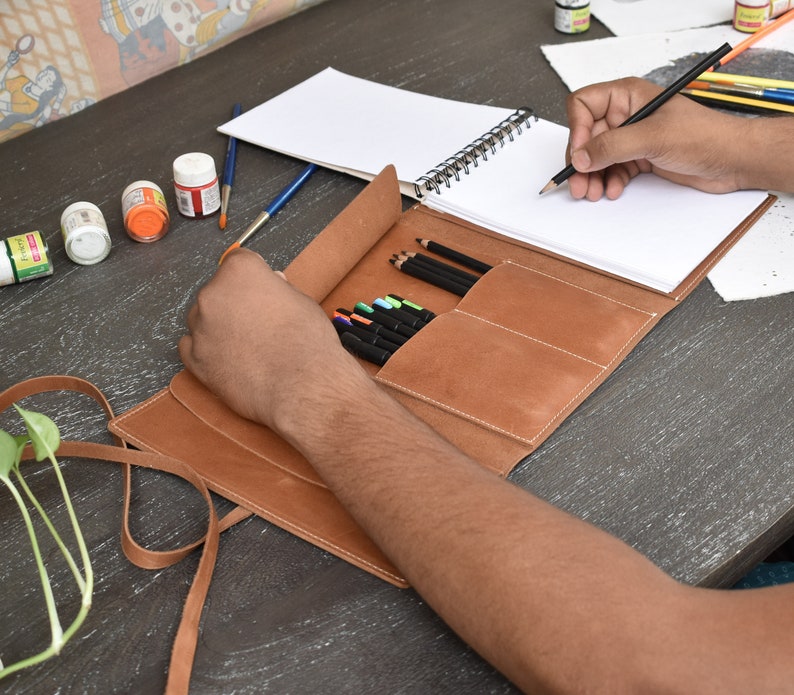 May include: A tan leather roll-up organizer featuring multiple interior pockets for pencils and pens, accompanied by a spiral-bound sketchbook with blank white pages. The organizer includes a long leather strap for securing the folded case.