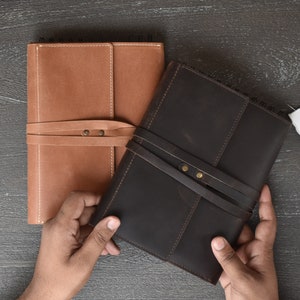 May include: Two rectangular notebooks with leather covers, one in tan and one in dark brown. Each notebook features a wrap-around leather strap secured with two metal rivets and a spiral binding along the top edge. The covers have visible stitching.