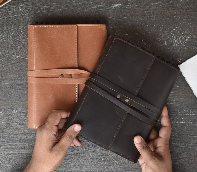 May include: Two rectangular notebooks with leather covers, one in tan and one in dark brown. Each notebook features a wrap-around leather strap secured with two metal rivets and a spiral binding along the top edge. The covers have visible stitching.