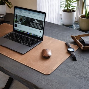 Pode incluir: Um tapete de mesa de couro marrom com um laptop, um mouse sem fio, um relógio de pulso e uma pequena bandeja de couro com uma carteira em uma mesa de madeira escura.