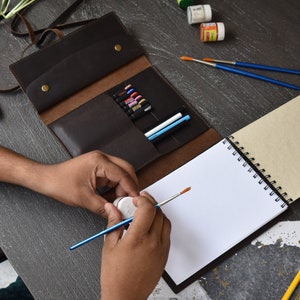 May include: A dark brown leather roll-up organizer case featuring multiple interior slots containing various pens and markers, secured with two brass-colored metal snap buttons, positioned next to a spiral-bound sketchbook and paintbrushes.