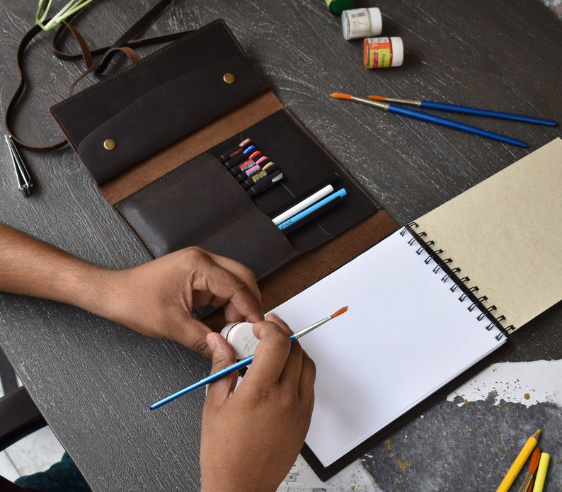 May include: A dark brown leather roll-up organizer case featuring multiple interior slots containing various pens and markers, secured with two brass-colored metal snap buttons, positioned next to a spiral-bound sketchbook and paintbrushes.