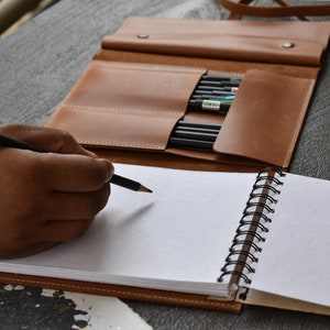 May include: A tan leather pencil roll with multiple slots containing black pencils, positioned behind a spiral-bound notebook with blank white pages. A black pencil is held in position over the paper, ready for writing or drawing on the page.
