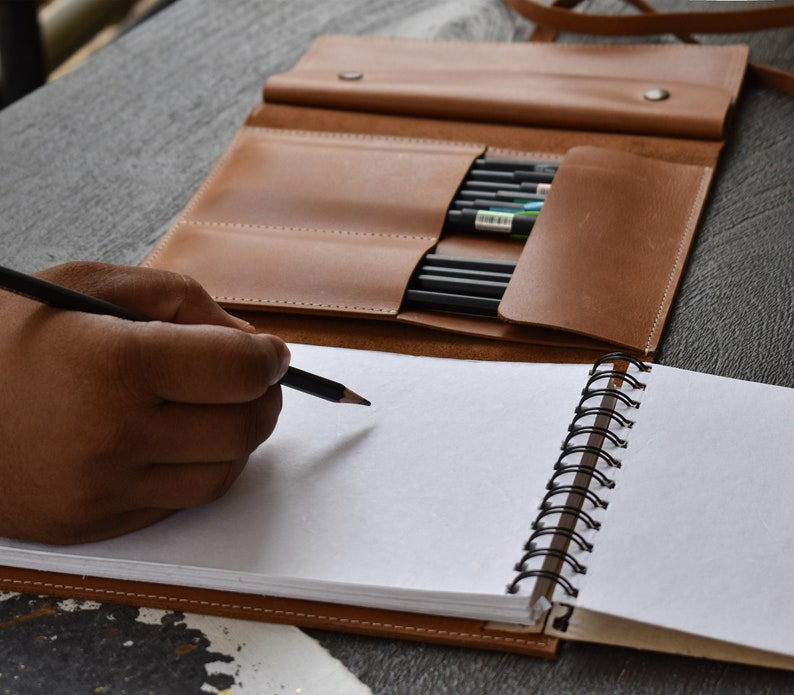 May include: A tan leather pencil roll with multiple slots containing black pencils, positioned behind a spiral-bound notebook with blank white pages. A black pencil is held in position over the paper, ready for writing or drawing on the page.