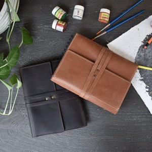 May include: Two rectangular leather-bound journals, one in dark brown and one in light tan, each featuring a wrap-around leather strap closure secured with two metal rivets. The journals are positioned on a dark surface alongside small paint pots.