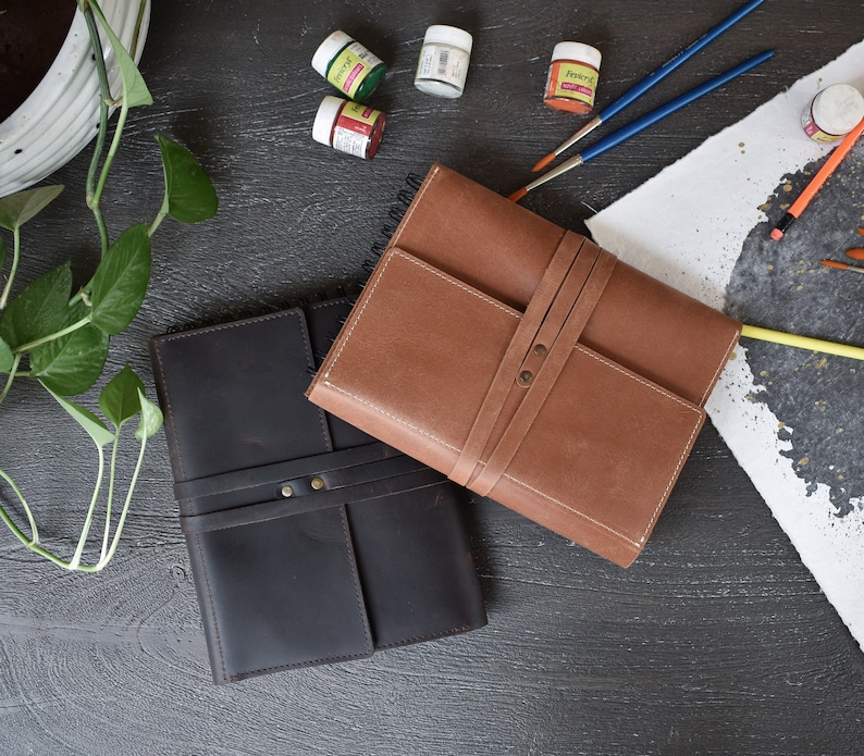 May include: Two rectangular leather-bound journals, one in dark brown and one in light tan, each featuring a wrap-around leather strap closure secured with two metal rivets. The journals are positioned on a dark surface alongside small paint pots.
