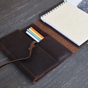 May include: A dark brown leather notebook cover featuring a wrap-around strap closure, a spiral-bound notebook with light beige pages, and an interior pocket containing four colored pens in white, blue, yellow, and orange, all stitched together.
