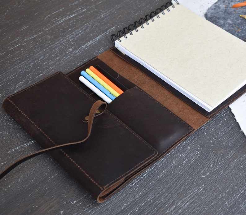 May include: A dark brown leather notebook cover featuring a wrap-around strap closure, a spiral-bound notebook with light beige pages, and an interior pocket containing four colored pens in white, blue, yellow, and orange, all stitched together.