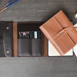 May include: A dark brown leather organizer featuring a flap with two brass snap buttons, multiple interior pockets holding various pens, and a separate light brown leather-bound notebook with a wrap-around strap closure and white paper pages.