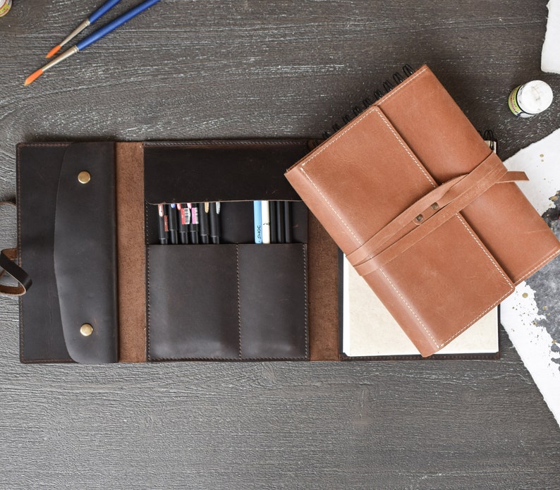 May include: A dark brown leather organizer featuring a flap with two brass snap buttons, multiple interior pockets holding various pens, and a separate light brown leather-bound notebook with a wrap-around strap closure and white paper pages.