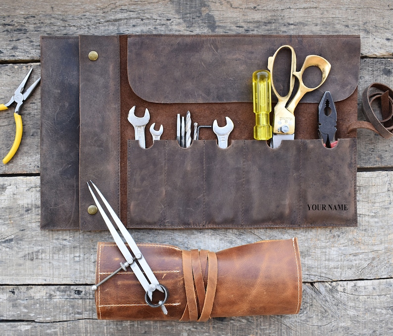 May include: A brown leather tool roll with multiple pockets for tools. The roll is rolled up and tied with a leather strap. The roll is open and shows a variety of tools, including a pair of pliers, a screwdriver, a wrench, and a pair of scissors. The roll is personalized with the text "YOUR NAME".