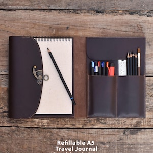 May include: A dark brown leather refillable A5 travel journal sketchbook cover with a metal latch closure. The interior features a spiral-bound notebook, a black pencil, and a storage section containing various pens, pencils, and a small ruler.