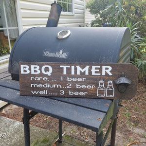 May include: A wooden sign with the text "BBQ TIMER" and a bottle opener. The sign has instructions for cooking meat based on the number of beers you drink. The sign is attached to a black grill.