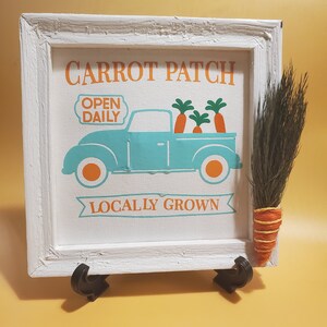 May include: White wooden sign with a blue truck carrying carrots and the text "Carrot Patch Open Daily Locally Grown".