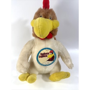 May include: A plush Foghorn Leghorn toy featuring a brown head, a bright red comb, and a large yellow beak. The body is off-white with yellow feet. A circular blue and cream tag on the chest displays an illustration of the character and the text Foghorn Leghorn.