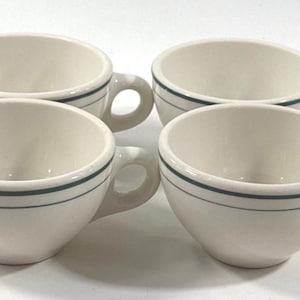 May include: Four off-white ceramic coffee cups with a dark green stripe near the rim and a handle. The cups are arranged in a square formation, showcasing their simple, classic design. Ideal for serving coffee or tea.