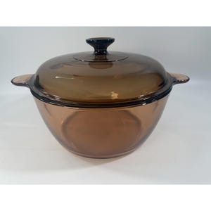 May include: A round, amber-colored glass casserole dish with a matching lid. The pot features two integrated side handles and a central knob on the lid. The glass is translucent, allowing light to pass through the brown-tinted material.