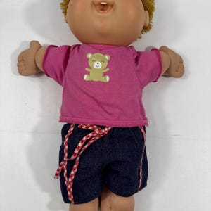 May include: A doll with light skin, blue eyes, and short blonde yarn hair. The doll wears a short-sleeved pink shirt featuring a small tan bear graphic on the chest and dark blue denim shorts with a red and white checkered drawstring tie.