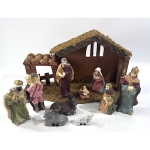 May include: A nativity set featuring a wooden stable with a moss-covered roof and eleven painted ceramic figurines. The set includes figures of Joseph, Mary, the infant Jesus in a manger, three wise men, a shepherd with a lamb, a donkey, and a sheep.