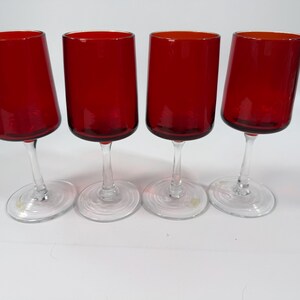 May include: A set of four matching wine glasses featuring deep red bowls and clear glass stems with circular bases. Each glass has a cylindrical bowl shape with a smooth finish, standing upright on a flat surface to display the vibrant red color.