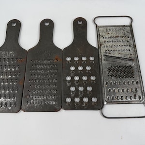 May include: Four vintage metal graters. Three graters have a paddle shape with a hole for hanging. The fourth grater is rectangular with a handle. All graters show signs of age and use, with a dark, aged metal finish.