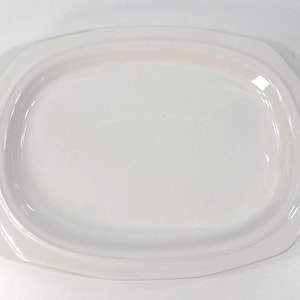 May include: A solid white ceramic serving platter with an oval-shaped center and a wide, flat rim featuring slightly angled corners. The surface is smooth and glossy, reflecting light across the entire piece, which has a clean, simple design.