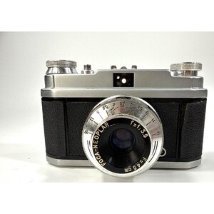 May include: A vintage Foca-Neoplar camera with a silver metal body and black textured grip. The lens is marked with F=4.5 cm and f=1:3.5. The top features two silver adjustment dials and a central viewfinder. The design is compact and rectangular.