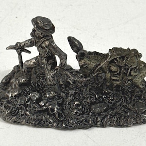 May include: A silver-toned metal figurine depicting a prospector wearing a hat and holding a pickaxe, leading a pack donkey loaded with supplies. The base features a textured ground surface with a small animal skull resting near the prospector.