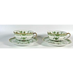May include: Two white porcelain teacups and matching saucers featuring a delicate green floral pattern and gold trim along the rims. Each cup has a curved handle and sits on a scalloped saucer, both decorated with intricate green botanical motifs.