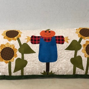 May include: A long, rectangular pillow with a white background and a patchwork border. The pillow features a scarecrow with a pumpkin head, a plaid shirt, and blue overalls. Two sunflowers with green stems and leaves are on either side of the scarecrow.