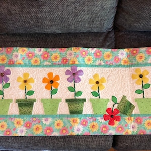 May include: A quilted wall hanging with a floral theme. It features six green flower pots with colorful flowers and a watering can. The background is a light blue fabric with a floral pattern.