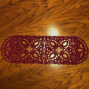 May include: A burgundy-colored, openwork lace table runner with an intricate floral and scroll design. The runner is oval-shaped and placed on a wooden table. The lace pattern is symmetrical and detailed, creating a decorative accent for home decor.