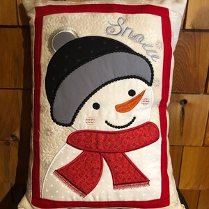 May include: A white pillow with a red border and a snowman applique. The snowman has a black hat, a gray scarf, and a red scarf with a white polka dot pattern. The word "Snowe" is embroidered on the pillow.