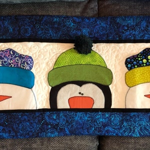 May include: A quilted wall hanging with three snowman faces. The snowmen are wearing colorful hats with pom-poms. The background is a blue and black paisley pattern.