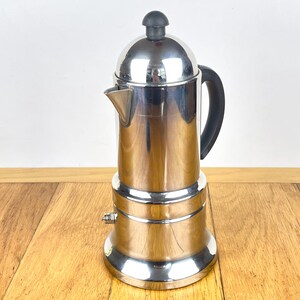 May include: A polished stainless steel stovetop espresso maker featuring a cylindrical body, a curved spout, and a black heat-resistant handle. The lid is topped with a small black knob, and the base is flared to provide stability on a burner.
