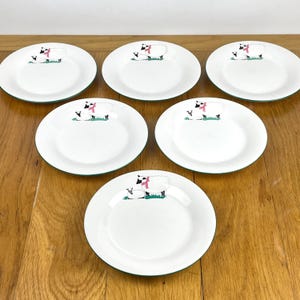 May include: Six white ceramic plates with a green rim. Each plate features a sheep illustration with a pink scarf and a smaller sheep on a patch of green grass. The plates are arranged on a wooden surface.