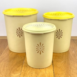 May include: Three vintage beige plastic canisters with yellow lids. Each canister has a brown floral design. The canisters are different sizes, with the largest one on the left and the smallest in the centre. The lids have a sunburst design.