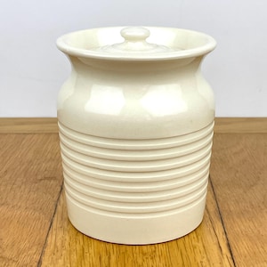 May include: Cream-coloured ceramic jar with a lid. The jar has a cylindrical shape with horizontal ridged detailing around the body. The lid has a small knob for easy removal. The jar sits on a wooden surface.