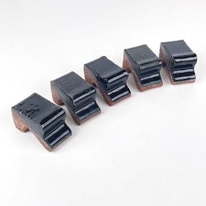 May include: Five small, L-shaped ceramic objects arranged in a row. Each piece features a glossy, dark black glazed top surface that contrasts with the exposed, unglazed reddish-brown clay base. The objects have a stepped, angular profile.