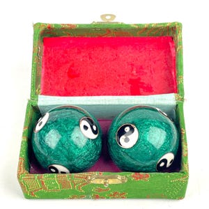 May include: Two spherical green meditation balls with a metallic finish and black and white yin-yang symbols, housed in a rectangular green fabric-covered box with a bright red interior lining and a gold-toned metal latch closure.