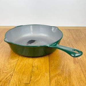 May include: A green cast iron frying pan with a long handle and a grey interior. The pan is on a wooden surface. The handle has a hole for hanging.
