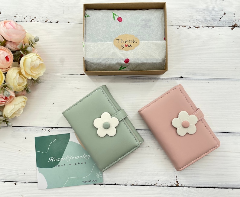Pu&ograve; includere: Due piccoli portafogli con disegni floreali. I portafogli sono realizzati in similpelle morbida. Un portafoglio &egrave; verde con un fiore bianco e l'altro &egrave; rosa con un fiore bianco. I portafogli sono su una superficie di legno bianco. Il testo "Hazel Jewelry Best Wishes Thank You" &egrave; su una carta in primo piano.