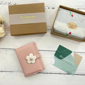 Pu&ograve; includere: Un portafoglio in similpelle rosa con un motivo a fiore bianco. Il portafoglio &egrave; confezionato in una scatola marrone con un biglietto di ringraziamento. Il biglietto dice "Hazel Jewelry Thank You".