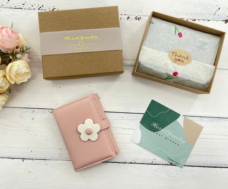 Pu&ograve; includere: Un portafoglio in similpelle rosa con un motivo a fiore bianco. Il portafoglio &egrave; confezionato in una scatola marrone con un biglietto di ringraziamento. Il biglietto dice "Hazel Jewelry Thank You".
