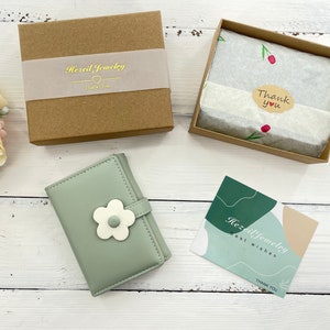 Pu&ograve; includere: Un portafoglio in pelle verde con un motivo a fiore bianco. Il portafoglio &egrave; chiuso e ha una piccola scatola marrone con il testo "Hazel Jewelry" su di essa. C'&egrave; anche un piccolo biglietto con il testo "Hazel Jewelry Best Wishes Thank You".