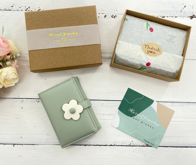 Pu&ograve; includere: Un portafoglio in pelle verde con un motivo a fiore bianco. Il portafoglio &egrave; chiuso e ha una piccola scatola marrone con il testo "Hazel Jewelry" su di essa. C'&egrave; anche un piccolo biglietto con il testo "Hazel Jewelry Best Wishes Thank You".
