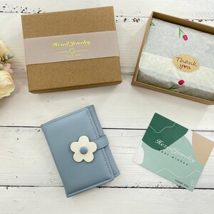 Pu&ograve; includere: Un portafoglio blu chiaro con un motivo a fiore bianco. Il portafoglio &egrave; chiuso e ha un piccolo lembo con una chiusura a bottone. Il portafoglio &egrave; su una superficie di legno bianco. L'immagine include anche un piccolo biglietto di ringraziamento con il testo "Hazel Jewelry Best Wishes Thank You" e una scatola marrone con il testo "Hazel Jewelry Thank You".