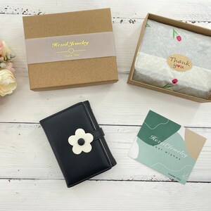 Pu&ograve; includere: Un portafoglio in pelle nera con un motivo a fiore bianco e il testo "Hezi Jewelry" su un biglietto bianco. Il portafoglio &egrave; su una superficie di legno bianco. Il biglietto dice anche "Auguri" e "Grazie".